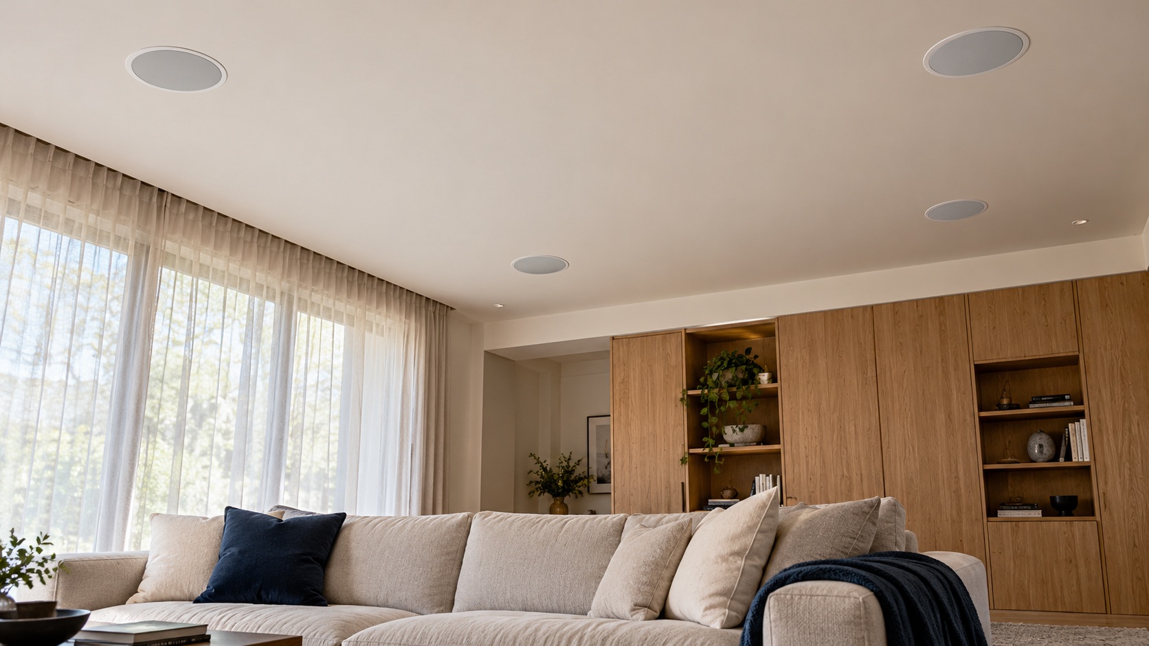 An illustrated living room seen from the sofa, with four round white speakers set flush into the ceiling.
