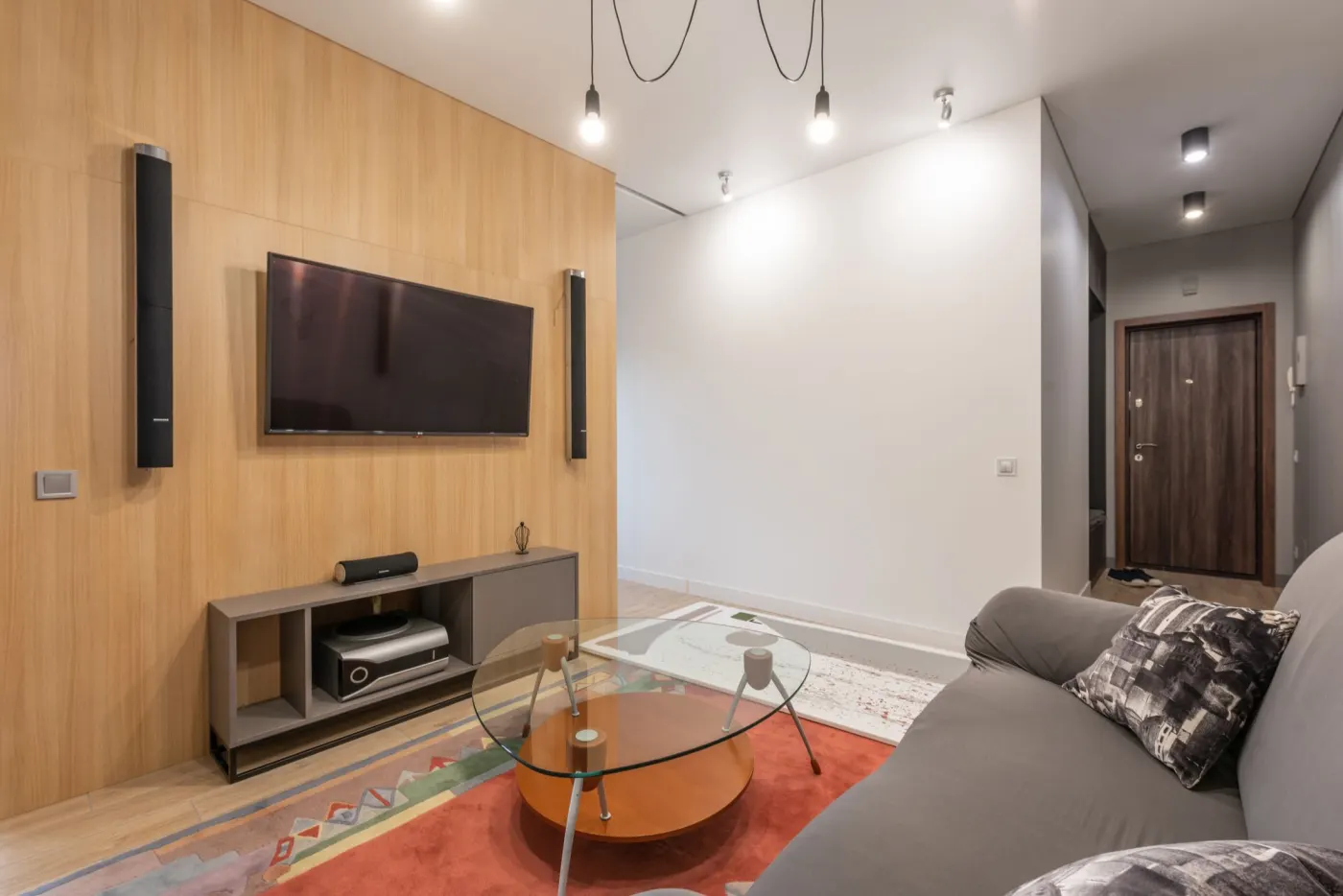 A living room with a wall-mounted TV between two slim tower speakers, a low media cabinet and a grey sofa.