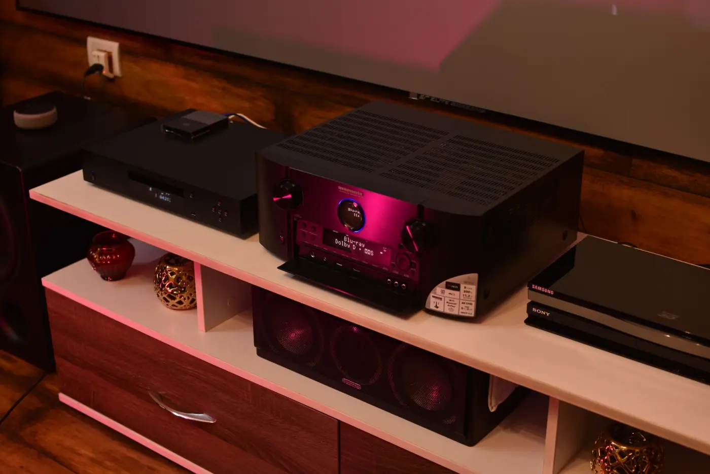 An AV receiver with a lit display on a white shelf, a disc player beside it and a centre speaker on the shelf below.