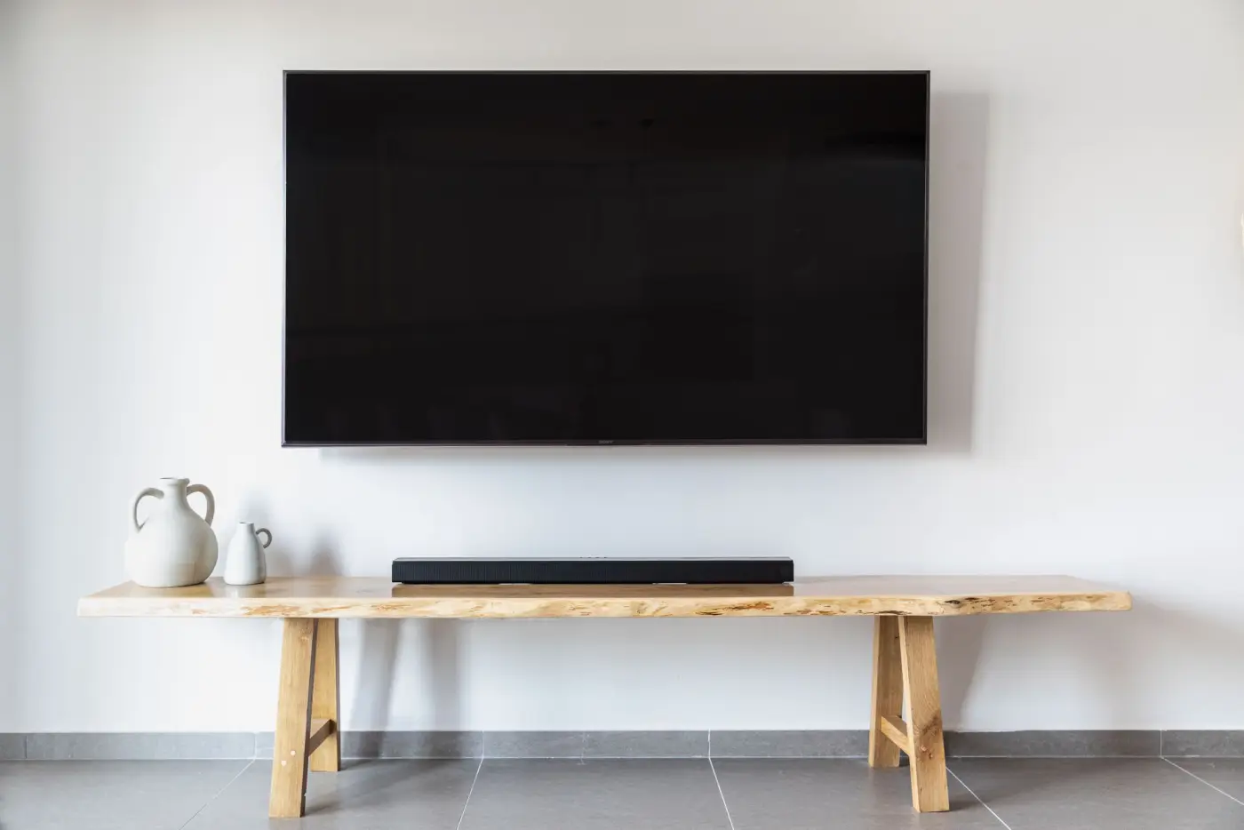 A wall-mounted television above a long wooden console with a slim soundbar and two white vases.