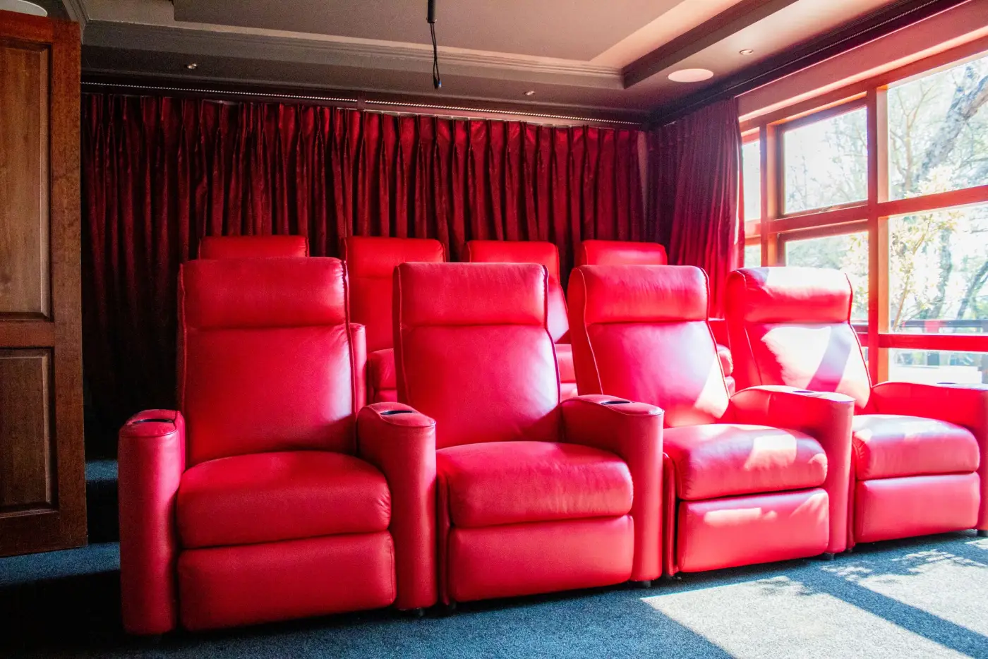 A home theatre room with two rows of red leather recliners, red curtains and a large window to one side.